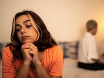 A couple sits apart on a bed, facing different directions. A woman, closer to the camera, looks pensive.